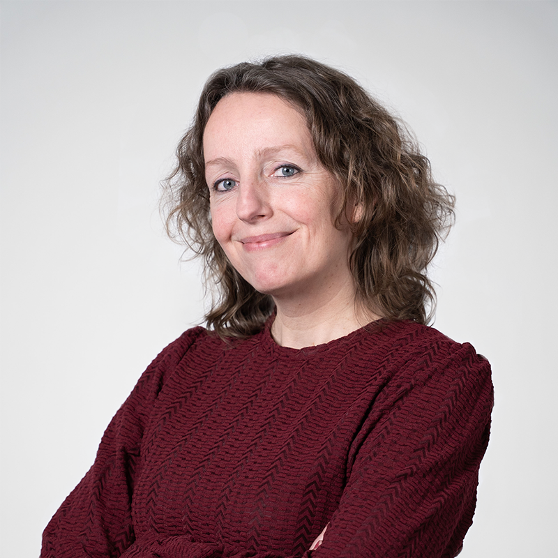 Portret van Marieke. Vrouw met krullen haar en een bordeaux rode trui. Ze kijkt vriendelijk.