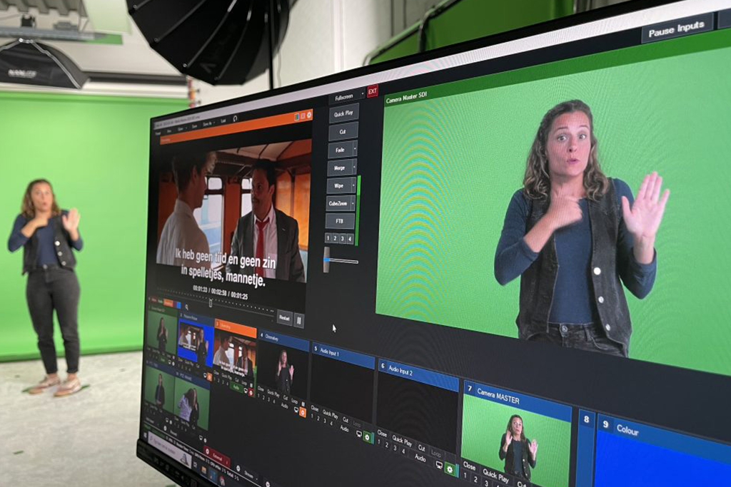 Monitor met videoproductiesoftware en een gebarentolk voor een groen scherm in een studio.